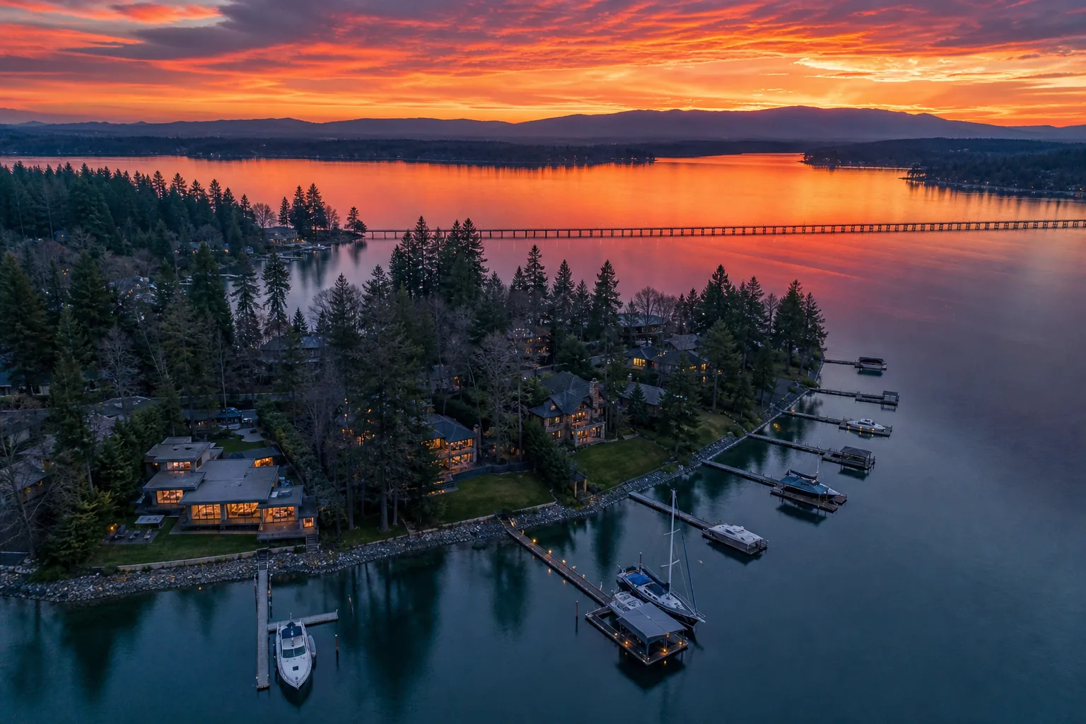 Aerial illustration at sunset of a wooded lakefront point lined with contemporary waterfront houses and long rows of private moorage, a low floating bridge crossing the still water beyond and banded orange cloud over distant hills